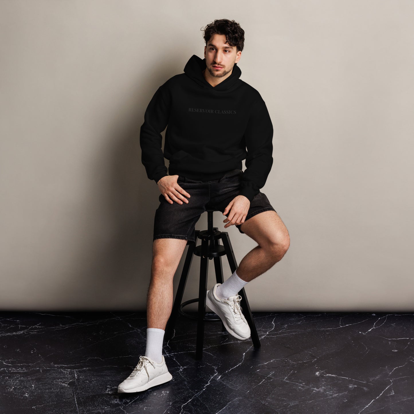 Reservoir Classics Unisex Men and Womens hoodie in black with no drawstring. A vintage, classic vibe streetwear style clothing line. Man sitting on stool to model